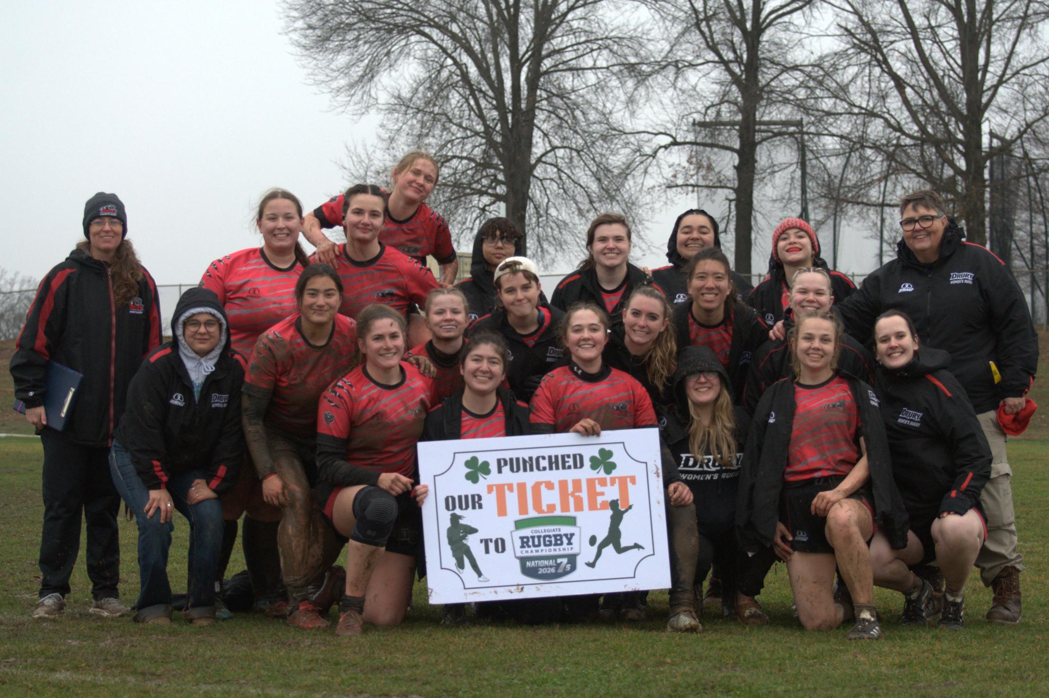 Drury University's Women's Rugby team holding a large sign, kneeling on the grass during a foggy spring day. Two trees behind them.