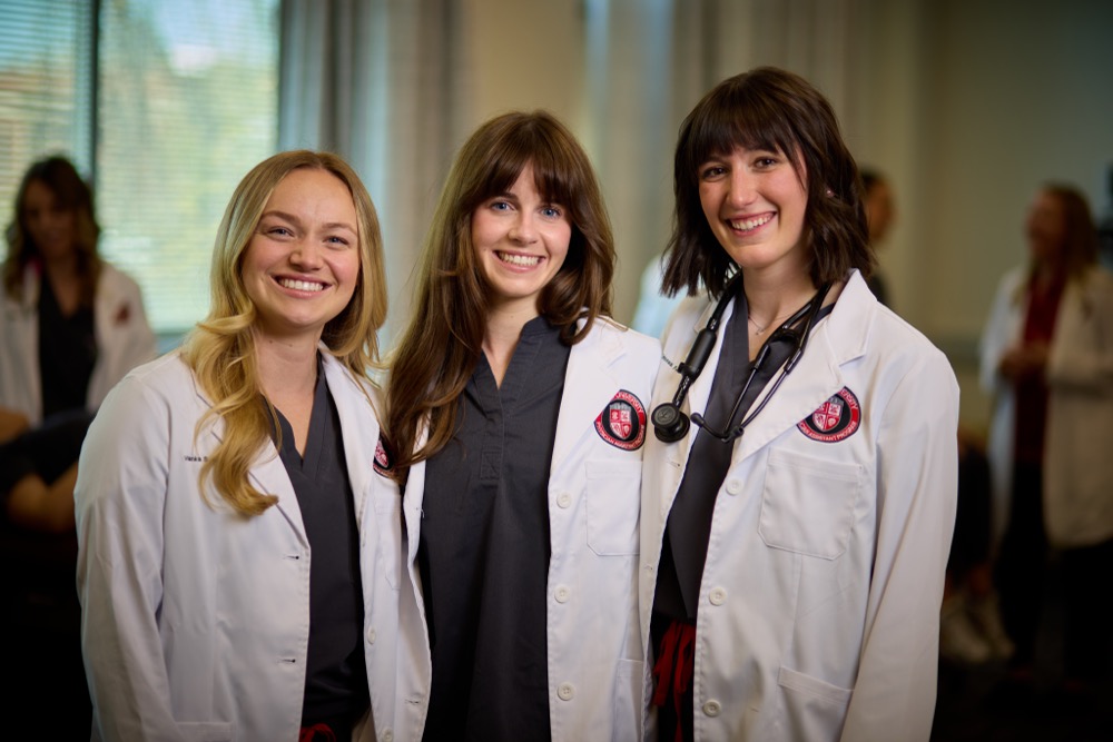 Three female Drury PA students smiling, and looking at the camera. They are all 3 wearing their white coat uniforms.