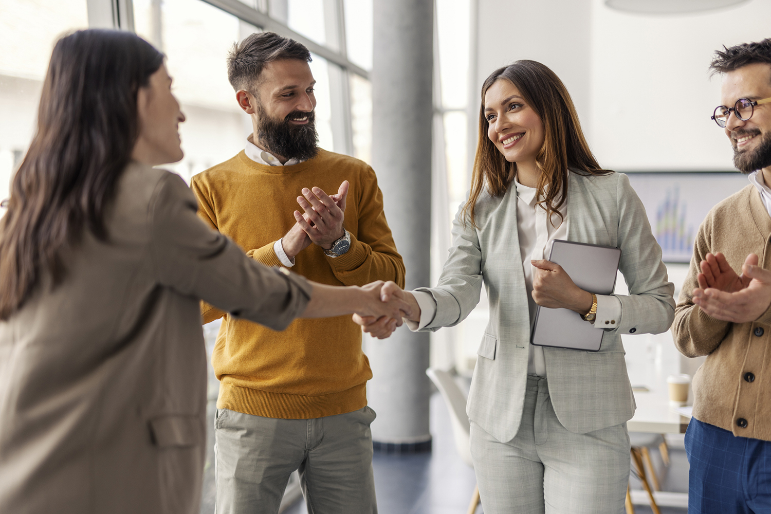 a business woman shaking hands with another woman. both are wearing blazers. In between them is a bearded man with a yellow sweater. on the right side is a man standing nearby smiling.