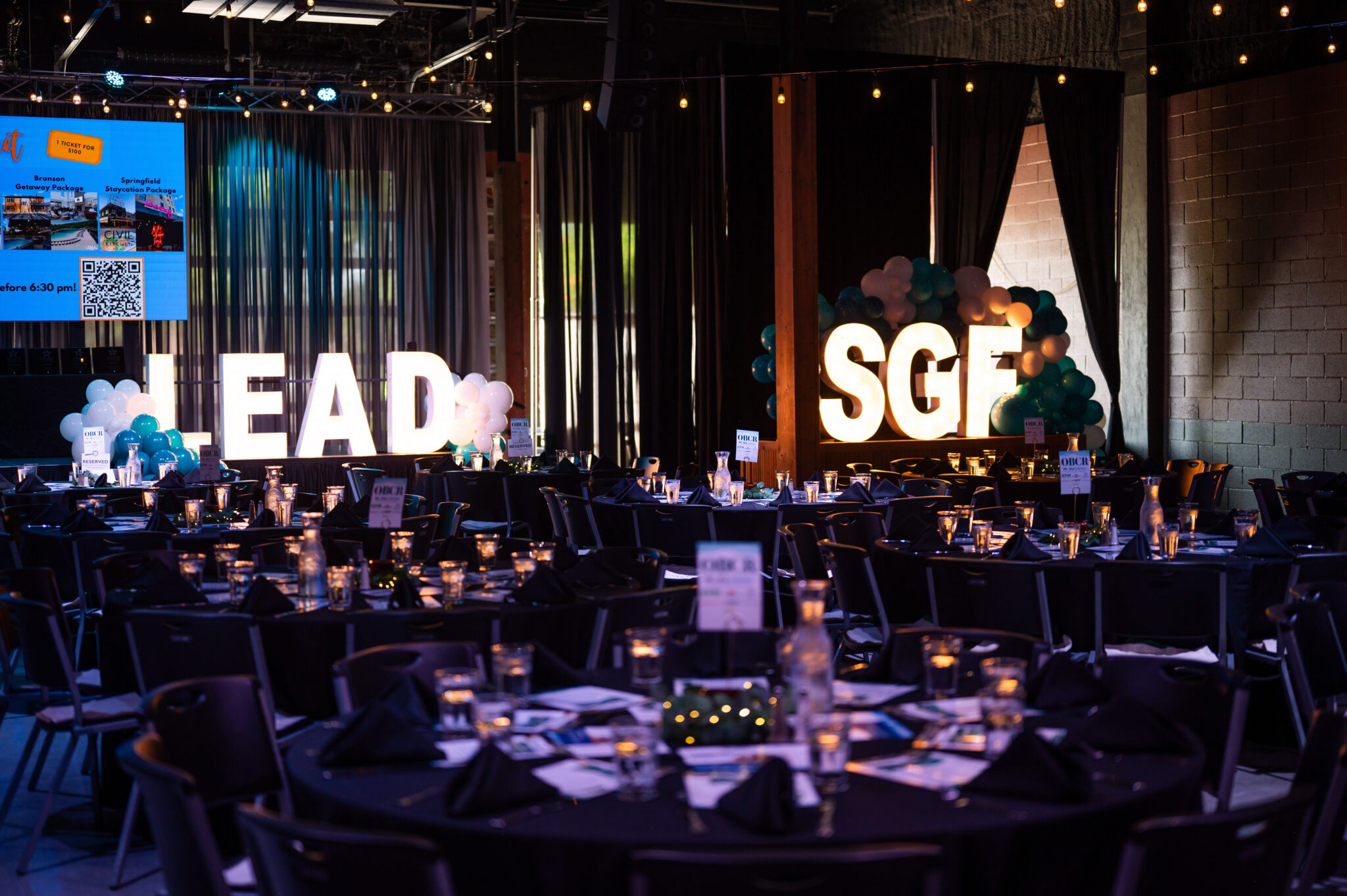 An image portraying a dimly lit ballroom with a set dinner table, and in the background a large LED sign with letters reading LEAD SGF, for a Leadership Springfield gala dinner event.