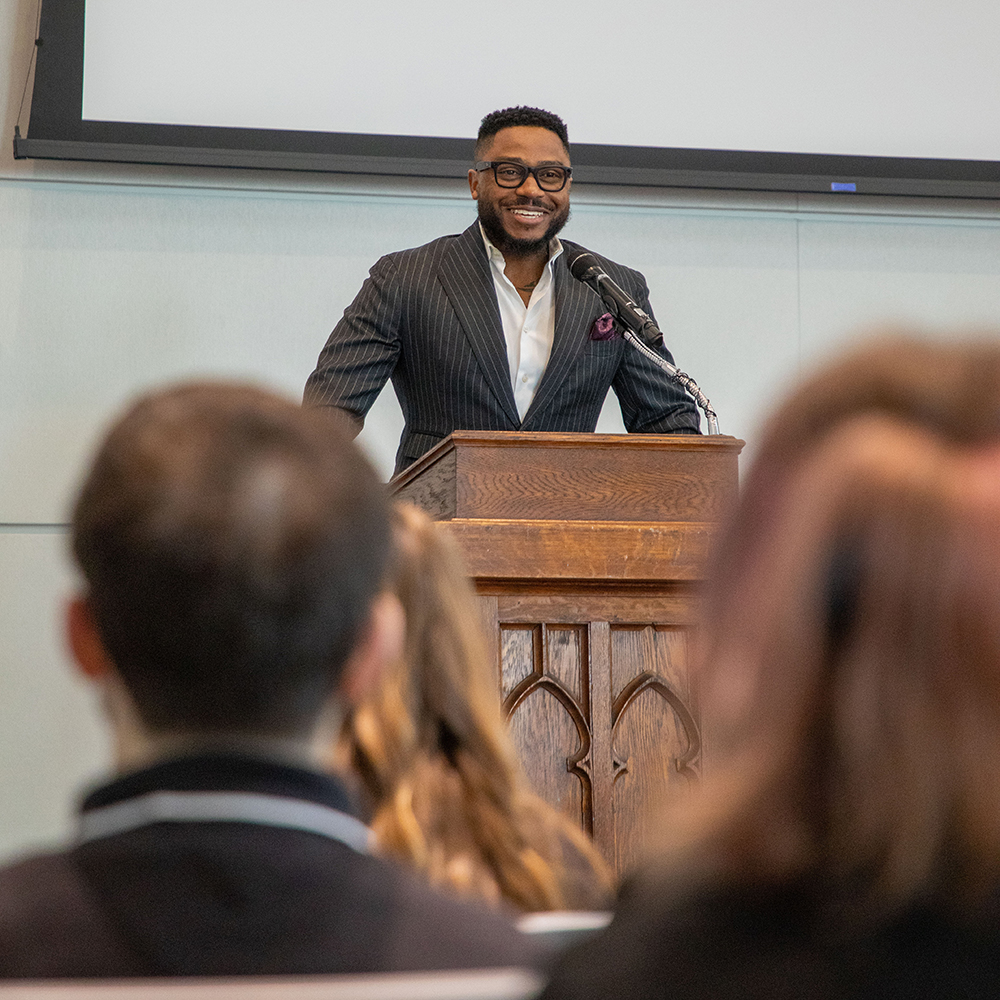 african american man wearing a suit, standing at a podium, looking at the camera and smiling in front of an audience