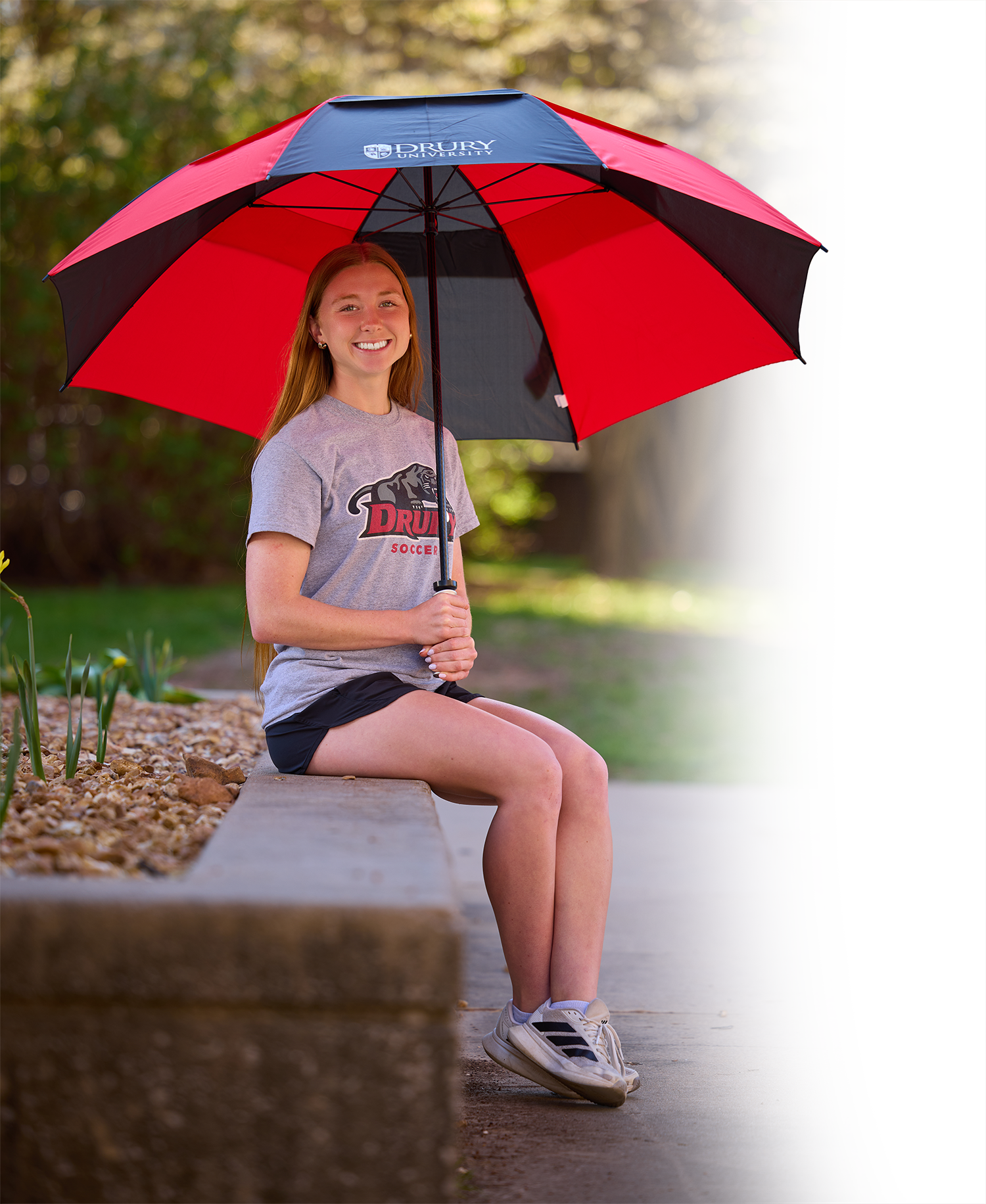 a young caucasian woman sitting on a concrete ledge, holding a black and red Drury-branded umbrella, looking at the camera and smiling