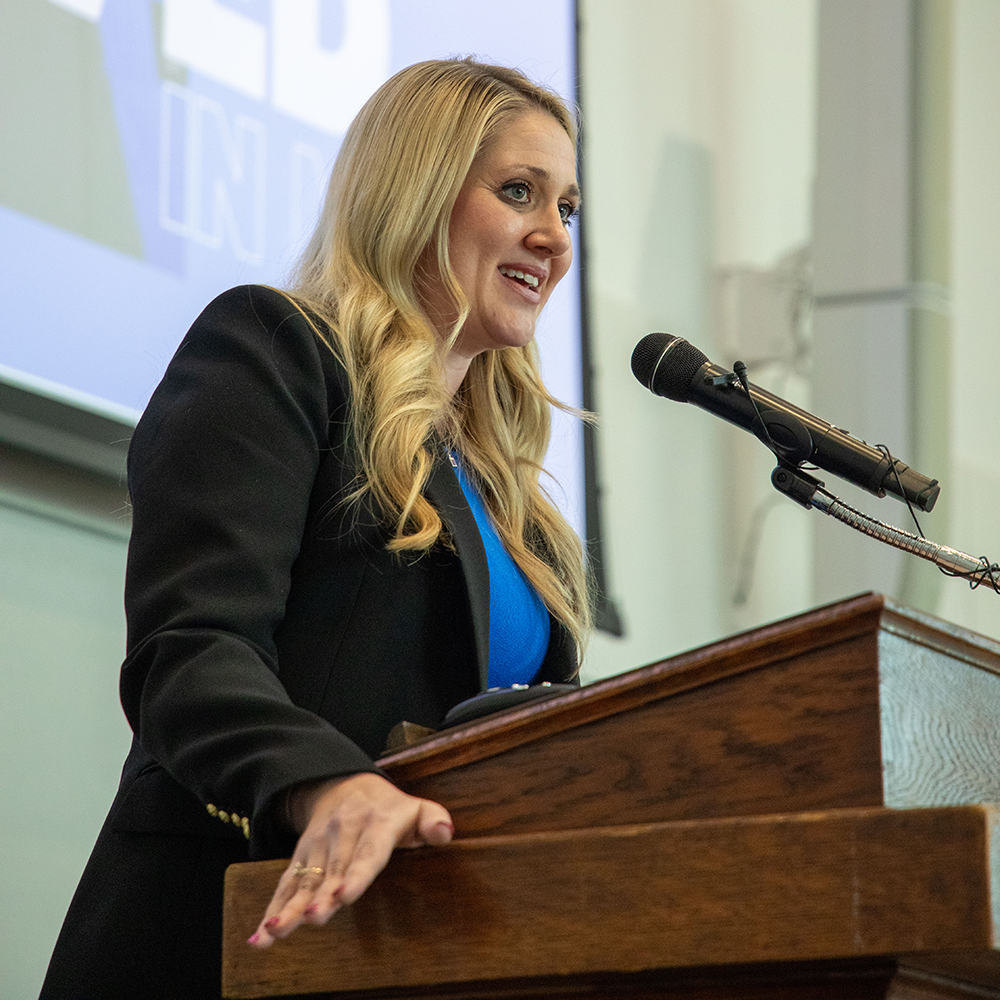 a caucasian woman with blonde hair standing at a podium, smiling and speaking