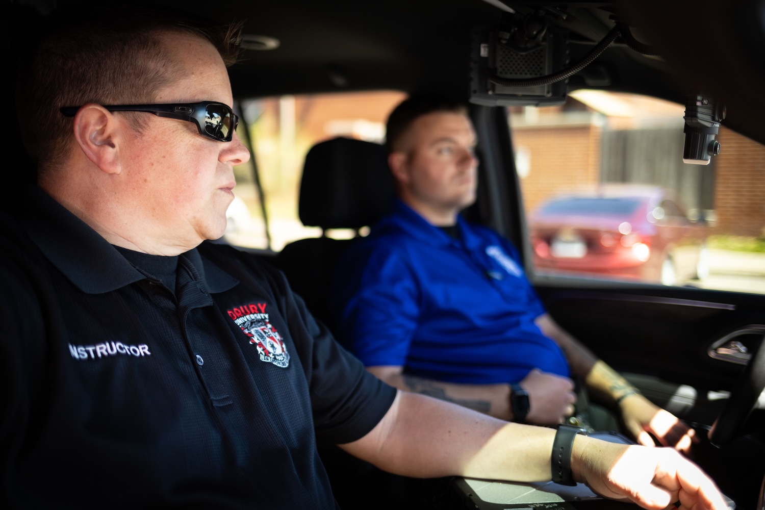 two white males, sitting in a vehicle, one wearing sunglasses and a black shirt, the other wearing a blue polo