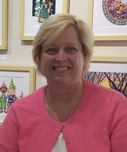 caucasian, middle aged woman with blonde short hair, wearing a pink blouse and white shirt. behind her is a wall of framed art pieces