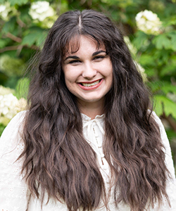 photograph of a smiling, young caucasian woman with brown eyes, long wavy brown hair, and a white dress.