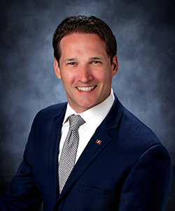 Studio photograph of a middle-aged brunette man wearing a dark suit and light blue tie, smiling at the camera