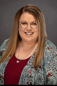 Studio portrait photograph of a middle aged woman with light brown hair and blonde highlights. She is wearing glasses, a necklace, a floral jacket, and a maroon shirt.